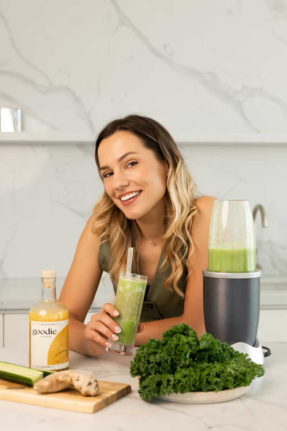 A woman smiling in her kitchen with a green smoothie and a bottle of Goodie