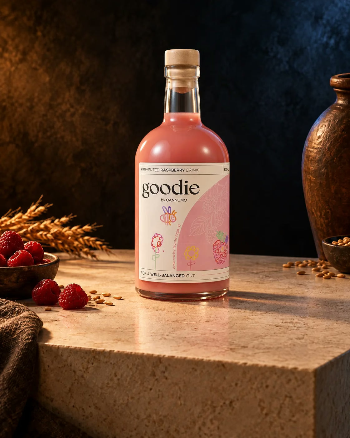 A bottle of Goodie Raspberry on a warm cream plinth with raspberries, grains and a fermentation vessel