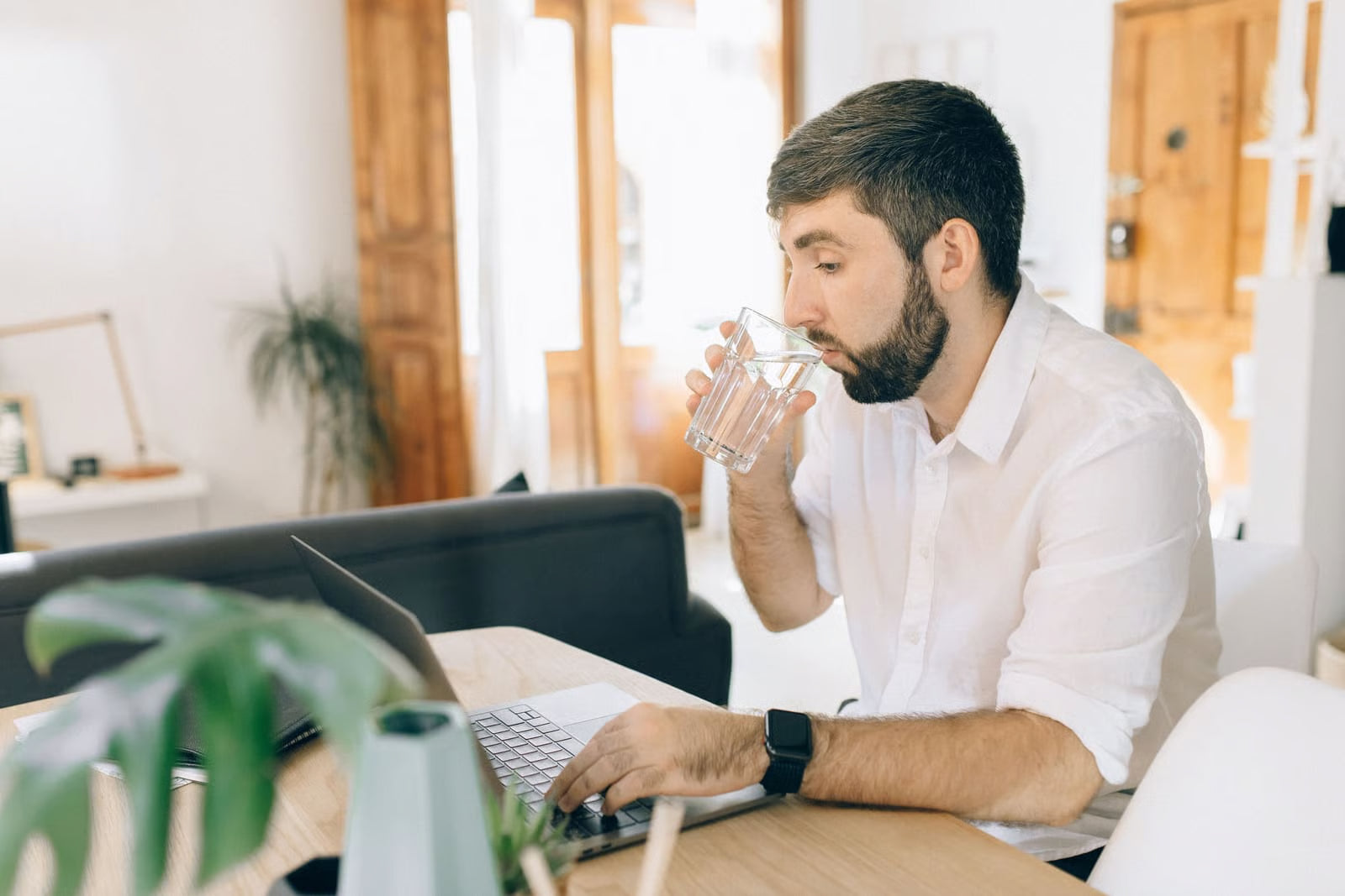 Man drinking a glass of water at his desk while working on a laptop